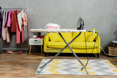housekeeping concept, stack of folded and clean clothes near iron on ironing board, modern living room, domestic life, laundry day, yellow sofa, domestic chores  clipart