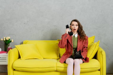 Telefon görüşmesi, dalgalı saçlı çekici bir kadın sarı koltukta oturuyor, ev hanımı retro telefonda konuşuyor, oyuncak bebek gibi davranıyor, kameraya bakıyor, ağzı açık, şok olmuş, gri duvarlı. 