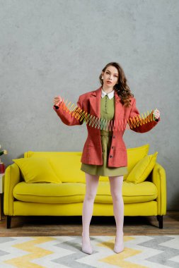 young brunette woman posing like a doll and playing with rainbow slinky, looking at camera, modern living room with yellow couch, childish, vintage, nostalgia, colorful toy, leisure and fun  clipart