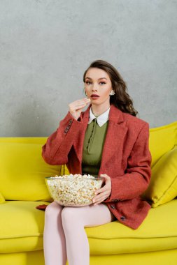 pretty girl acting like a doll, concept photography, young woman with brunette wavy hair eating popcorn, holding bowl, salty snack, home entertainment, sitting on comfortable yellow sofa  clipart
