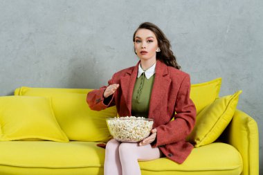 pretty girl acting like a doll, concept photography, young woman with wavy holding bowl with popcorn, gesturing unnaturally, salty snack, home entertainment, sitting on comfortable yellow sofa  clipart