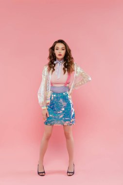 beautiful woman with wavy hair posing like a doll on pink background, conceptual photography, doll pose, girly outfit, model in skirt with sequins and transparent jacket standing with hand on hip clipart