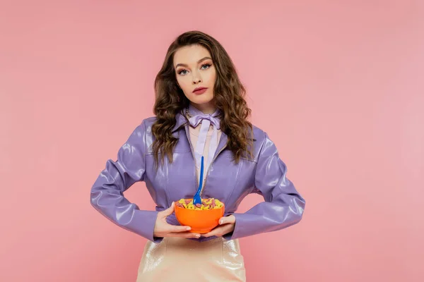 stock image concept photography, brunette woman with wavy hair pretending to be a doll, holding bowl with corn flakes and spoon, tasty breakfast, posing on pink background, stylish purple jacket