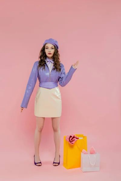 stock image fashion concept, doll-like, attractive young woman in beret standing near shopping bags on pink background, posing like a doll and looking at camera, trendy outfit, consumerism 