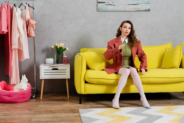 concept photography, posing like a doll, well dressed young woman with wavy hair sitting on yellow couch, gesturing, stylish house interior, vase with tulips, looking at camera, housewife lifestyle