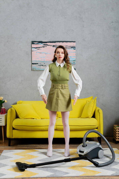 housekeeping concept, young woman with wavy hair standing on clean carpet near vacuum cleaner and yellow couch, gesturing and looking at camera, housewife in dress, domestic life, posing like a doll 