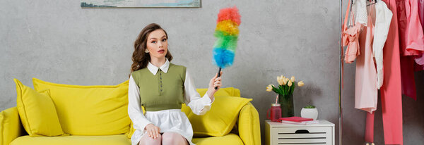 housekeeping concept, young woman with wavy hair sitting on couch and holding dust brush, housewife in dress and white tights looking at camera, domestic life, posing like a doll, banner 