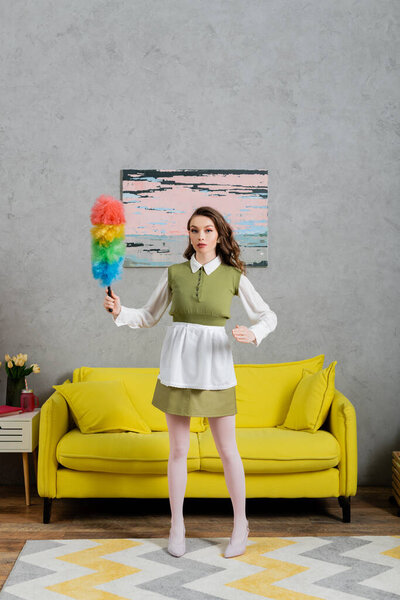 housekeeping concept, young woman with wavy hair standing and holding dust brush, housewife in dress and white tights looking at camera, gesturing unnaturally, domestic life, posing like a doll 