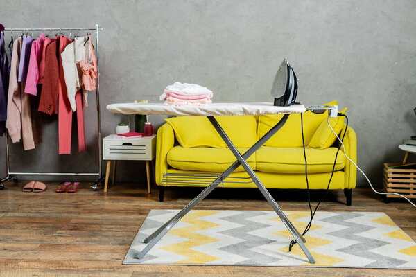 housekeeping concept, stack of folded and clean clothes near iron on ironing board, modern living room, domestic life, laundry day, yellow sofa, domestic chores 