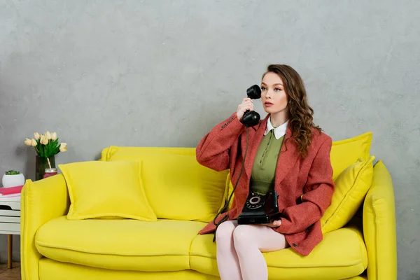 ヴィンテージビブ 黄色のソファに座って波状の髪の魅力的な女性 レトロな電話で話す主婦 人形のように振舞う 離れて見る 現代的なインテリア リビングルーム — ストック写真