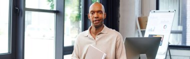 Myastthenia Gravis hastalığı olan koyu tenli bir adam. Dosyasını tutuyor ve ofisinde duruyor. Cesur bir Afro-Amerikan çalışanı kameraya, ekrana ve grafiklere bakıyor. 