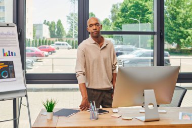 Ofis içi, ptosis sendromlu cesur Afro-Amerikan işadamı kameraya, miastenia gravis sendromuna, bilgisayar monitörüne, kırtasiyeye, masasında grafikler olan çizelgeye bakıyor.