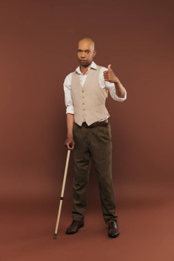 Myastthenia Gravis sendromlu Afro-Amerikan bir adam ayakta ve bastona yaslanıyor, kameraya bakıyor, kronik hastalığı olan koyu tenli cesur bir adam, kahverengi arka planda, 