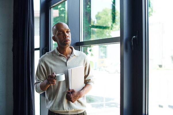 african american man with myasthenia gravis disease holding folder and cup of coffee, bold and dark skinned office worker with ptosis eye syndrome, inclusion, standing near windows  