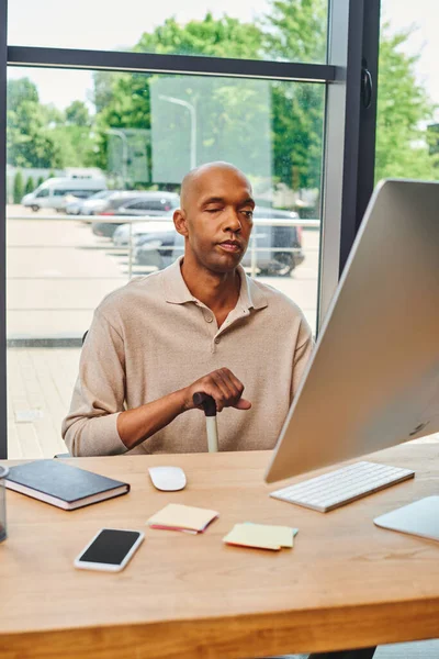inclusion, bold african american man with myasthenia gravis, dark skinned office worker sitting with walking cane and using computer, looking at monitor, keyboard and notebook on desk