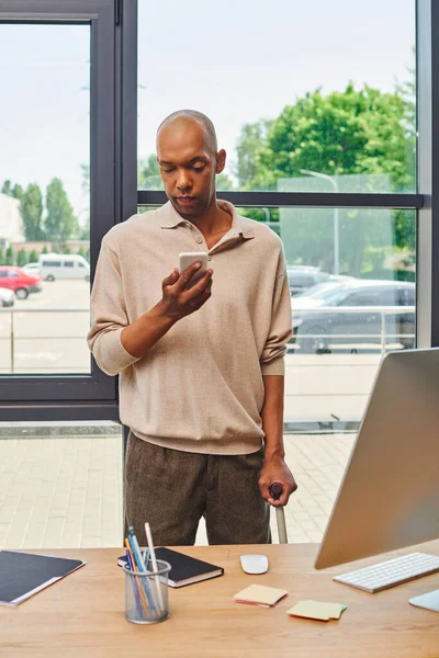 myasthenia gravis, bold african american businessman with eye syndrome holding smartphone and standing with walking cane near monitor, dark skinned office worker with ptosis syndrome, inclusion