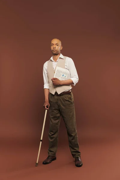 inclusion, bold african american man with myasthenia gravis syndrome, standing with newspaper and walking cane, dark skinned man with chronic disease on brown background