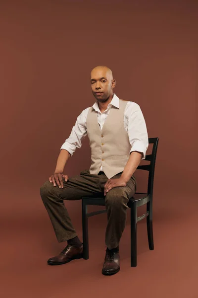 stock image inclusion, bold dark skinned man with myasthenia gravis syndrome, sitting on chair and looking at camera, african american man with chronic disease on brown background