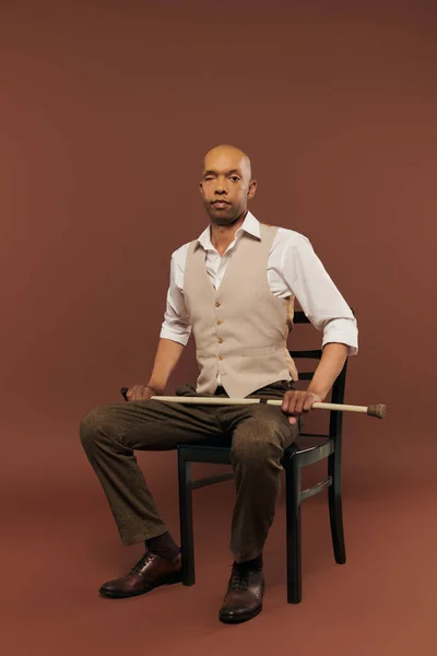 inclusion, bold african american man with myasthenia gravis syndrome, sitting on chair and looking at camera, dark skinned man holding walking cane on brown background, real people
