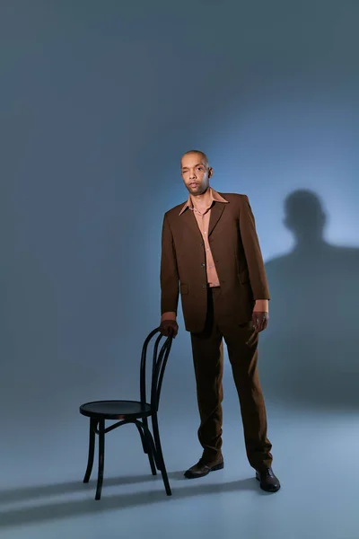 real people, bold african american man with myasthenia gravis syndrome standing with walking cane near chair on blue grey background, dark skinned person in suit, diversity and inclusion