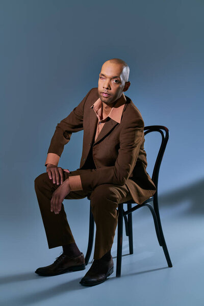 bold african american man with myasthenia gravis syndrome sitting on chair and looking at camera on blue background, dark skinned person in suit, diversity and inclusion, medical condition 
