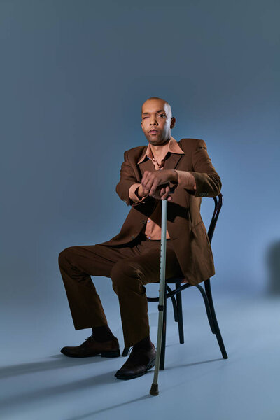 diversity and inclusion, african american man with myasthenia gravis syndrome sitting on chair and looking at camera on blue background, leaning on walking cane, difficulty walking 