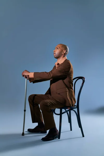 diversity and inclusion, physical impairment, african american man with myasthenia gravis sitting on chair and looking away on blue background, leaning on walking cane, difficulty walking