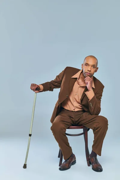 disability, full length of bold african american man with myasthenia gravis sitting on wooden chair on grey background, dark skinned person in suit leaning on walking cane, diversity and inclusion