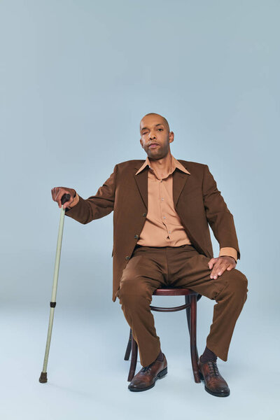diversity, full length of bold african american man with myasthenia gravis sitting on wooden chair on grey background, dark skinned person in suit leaning on walking cane, inclusion 