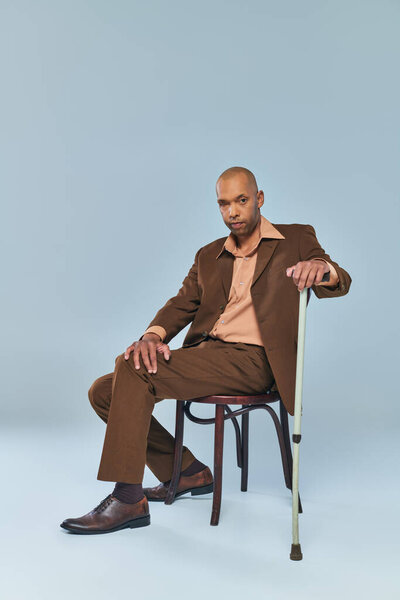 ethnic, full length of bold african american man with myasthenia gravis sitting on chair on grey background, dark skinned person in suit leaning on walking cane, diversity and inclusion 