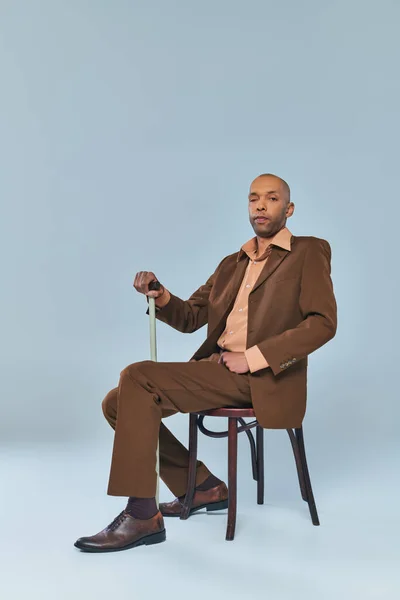 ptosis syndrome, full length of bold african american man with myasthenia gravis sitting on chair on grey background, dark skinned person in suit leaning on walking cane, diversity and inclusion