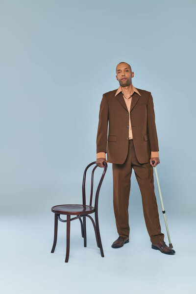 ptosis syndrome, full length of bold african american man with myasthenia gravis standing near chair on grey background, dark skinned person in suit leaning on walking cane, diversity and inclusion 