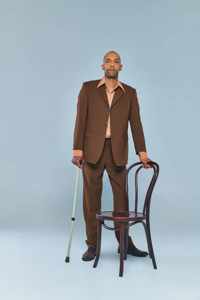 ptosis syndrome, full length of bold african american man with myasthenia gravis standing near chair on grey background, dark skinned person in suit with walking cane, diversity and inclusion