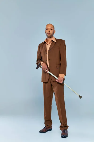 ptosis syndrome, full length of bold african american man with myasthenia gravis standing on grey background, dark skinned person in suit holding walking cane, diversity and inclusion