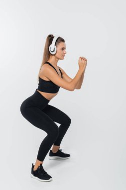 sportswoman in headphones and sportswear doing squats on white background, wellness concept clipart