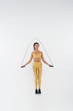 full length of brunette sportswoman in yellow activewear holding skipping rope and training on white clipart