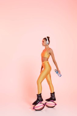 Kangoo zıplayan ayakkabılı ve kablosuz kulaklıklı bir kadın. Elinde pembe üzerine su dökülmüş spor şişesi tutuyor. 