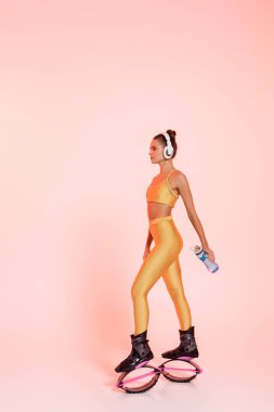 woman in kangoo jumping shoes and wireless headphones holding sports bottle with water on pink  clipart