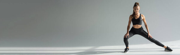 брюнетка спортсменка в спортивном бюстгальтере и леггинсах, смотрящая в камеру, упражняясь на сером фоне 