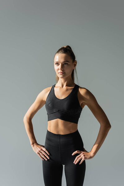 fit sportswoman in sports bra and leggings holding hands on hips and looking away isolated on grey 