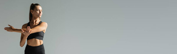 confident sportswoman in sports bra stretching arms and training isolated on grey, banner 