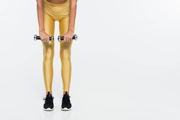 Cropped view of fit sportswoman in yellow leggings training with dumbbells on white background 