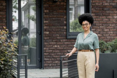 cheerful african american real estate agent smiling near fence of city cottage clipart