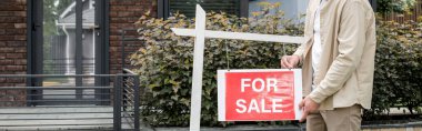 cropped view of property realtor hanging for sale signboard near contemporary city house, banner clipart