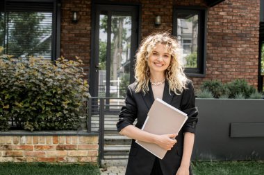Elinde belgelerle gezen neşeli sarışın emlakçı ve şehir caddesindeki evin yakınındaki kameraya bakıyor.