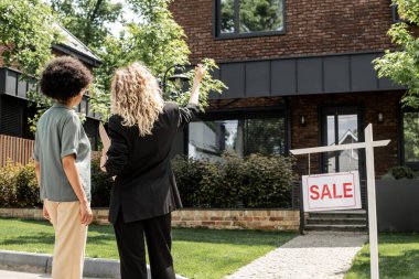 back view of blonde realtor pointing with hand, showing cottage for sale to african american woman clipart