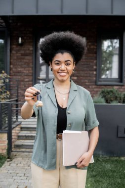 Mutlu ve başarılı Afrikalı Amerikalı emlak komisyoncusu elinde klasörle New Cottage 'ın yanında anahtar tutuyor.