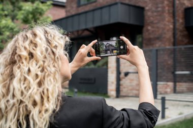 Sarışın emlak komisyoncusu cep telefonuyla şehir caddesinde çağdaş kulübenin fotoğrafını çekiyor.