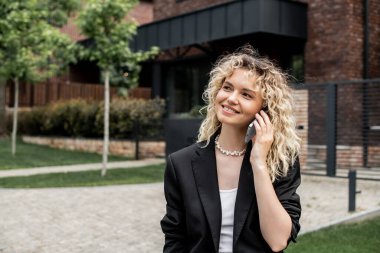 Güler yüzlü emlakçı şehir caddesindeki binanın yanında akıllı telefondan konuşurken başka tarafa bakıyor.