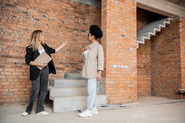 full length of real estate broker with folder showing unfinished interior to african american woman clipart